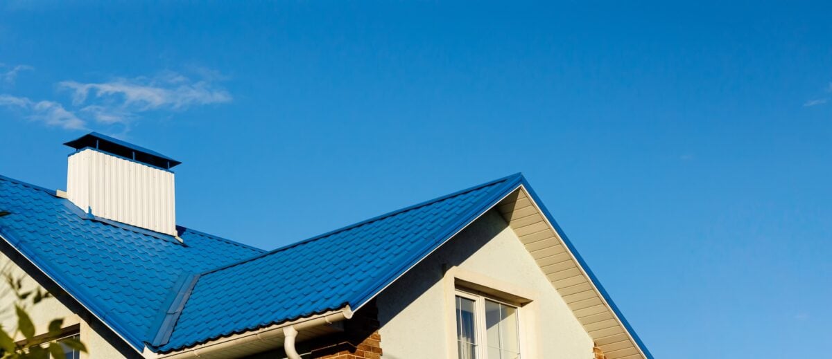 A roof of a house or cottage made of blue metal tiles with drains, slopes and chimney against the blue sky. Copy space