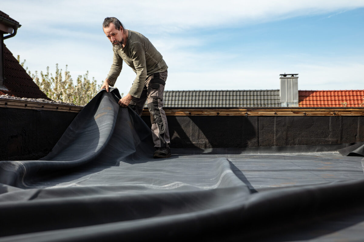 A craftsman rolls out a thick rubber tarpaulin on a flat roof to re-seal and repair the broken bitumen coating how long does a rubber roof last