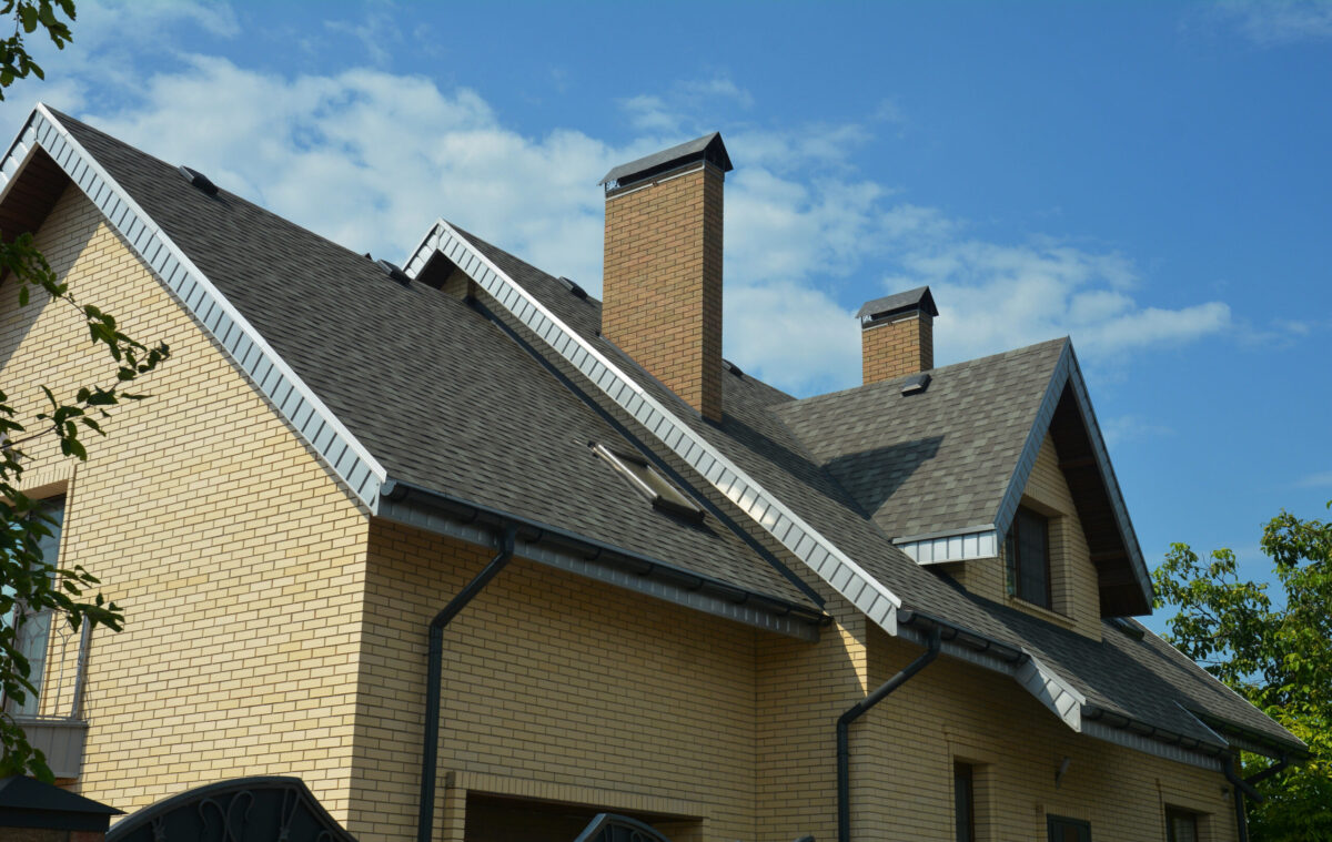 An asphalt shingled roof with skylights, attic windows, chimneys, fascias, and a roof gutter. A close-up of a gray double-pitched roof covered with dimensional asphalt roofing shingles.