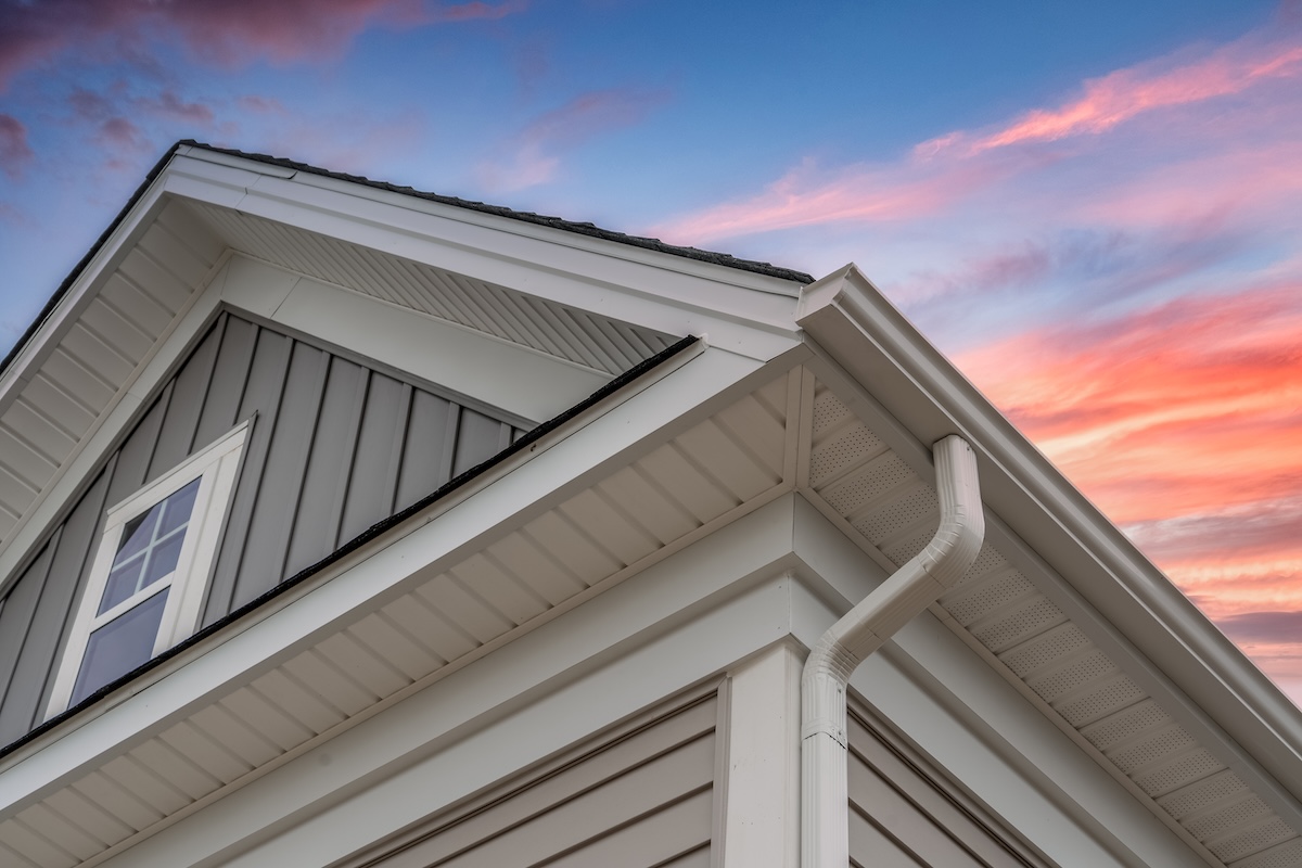 White frame gutter guard system, with gray horizontal and vertical vinyl siding fascia, drip edge, soffit, on a pitched roof attic at a luxury American single family home dramatic sunset sky what is a soffit