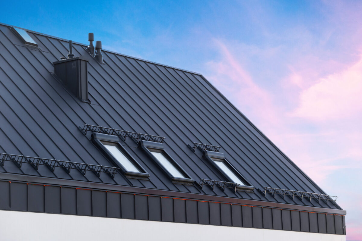 cost of metal roof vs shingle House with metal galvanized coating, chimney pipe, mansard windows and snow holding cornice on roof slope against sunset sky. Element of modern residential building with zinc rooftop sheets