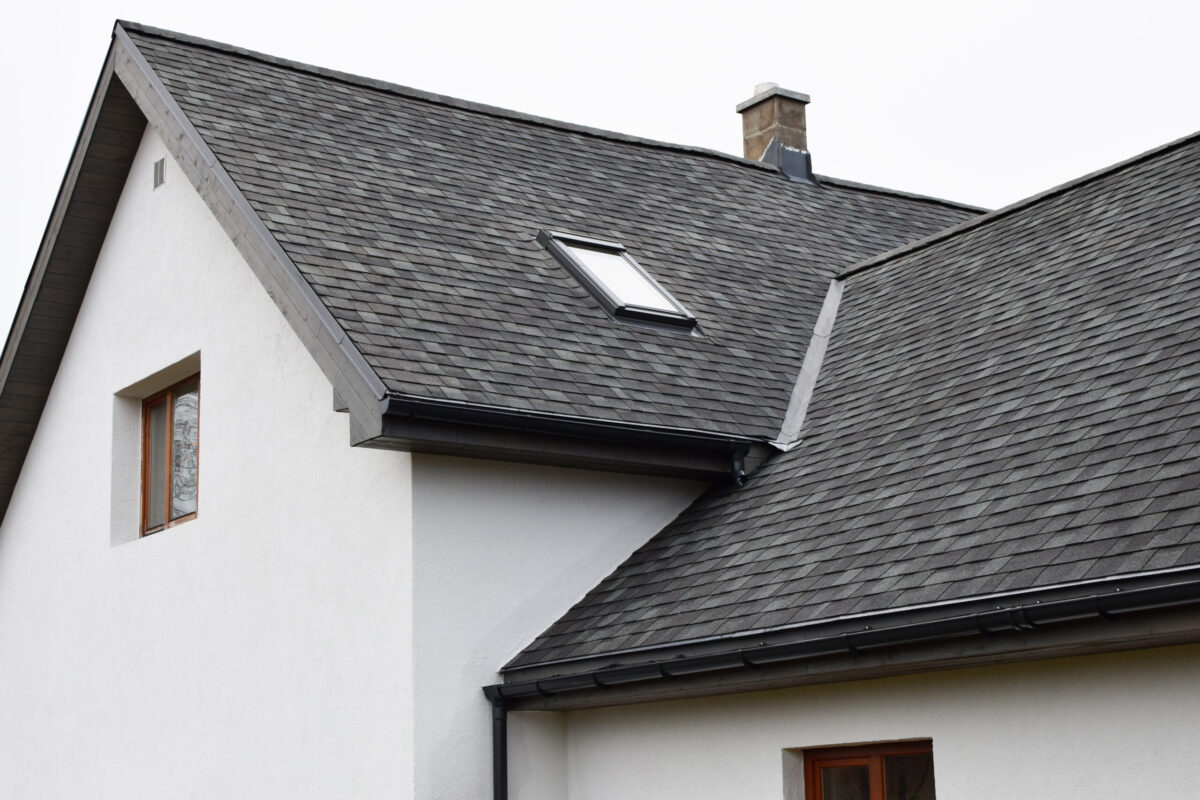 cost of metal roof vs shingle Part of white house with new wooden eaves, plastic rain gutter, window and chimney in grey shingles roof and brown window in facade.